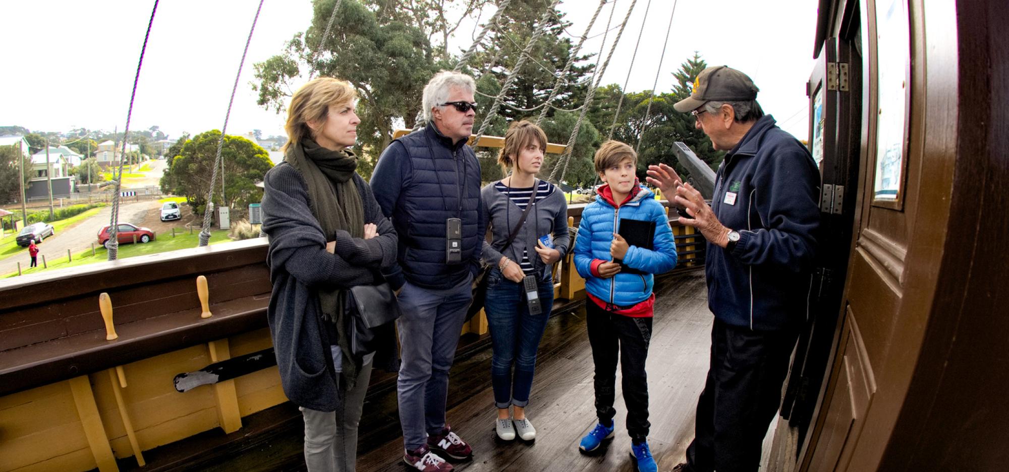 A slight fish-eye filtered image of a family of four wearing warm layers of clothing listening with interest to a person talking animatedly with his hands. They stand aboard the Brig Amity, an old ship, with a suburban street of Albany in the distance.