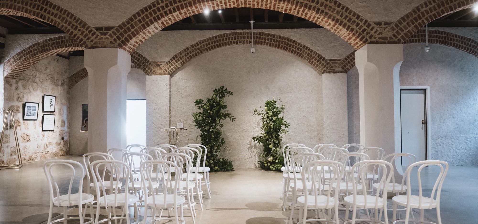 Chairs and flowers are arranged for a wedding in a large room with limestone walls and an arched ceiling