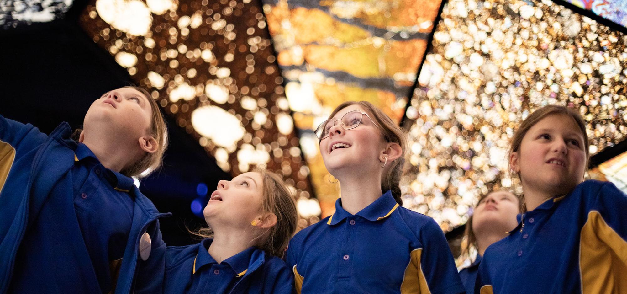 Four school children in blue uniforms stare up in awe at the colourful kaleidoscopic mineral tunnel at the WA Museum Boola Bardip