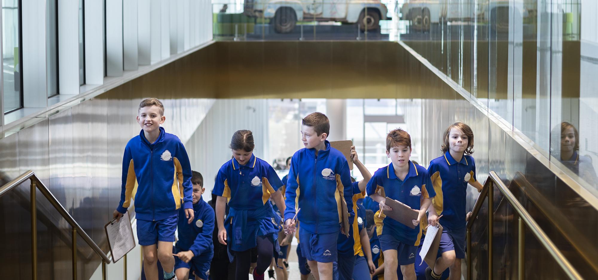 A large group of school children in blue school uniforms walk up a flight of stairs with terrazzo floors and gold fixtures with looks of excitement on their faces 