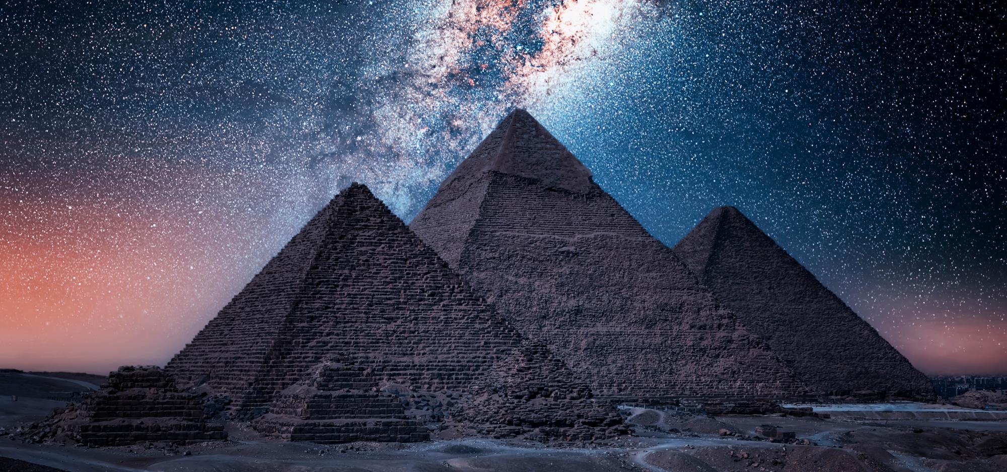 Three pyramids stand in darkness in a desert at night, backlit by the milkyway which shines vividly in the background in hues of orange, blue, purple and white, surrounded by hundreds of stars