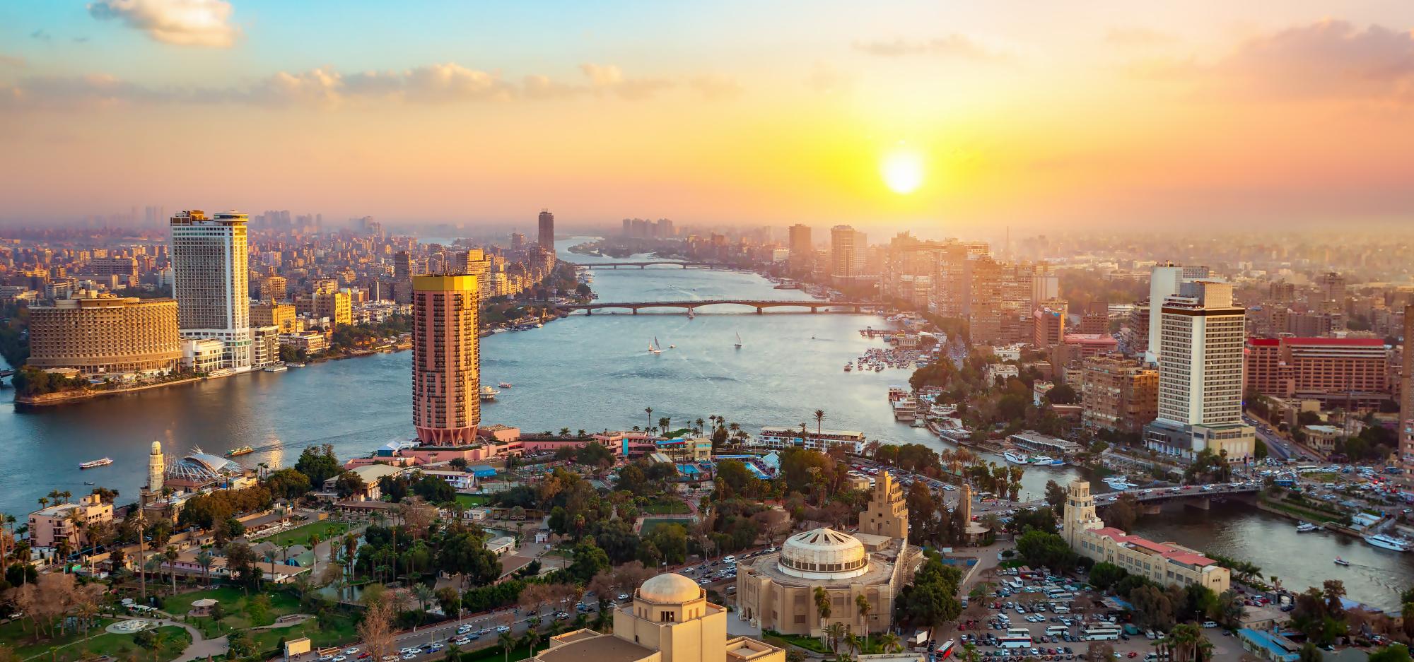 An idyllic panoramic view of the modern Cairo skyline, showing a sprawling city placed on either side of a large river. Under a bright yellow sunset, the towering skyscrapers and intricate temples appear to glow gold