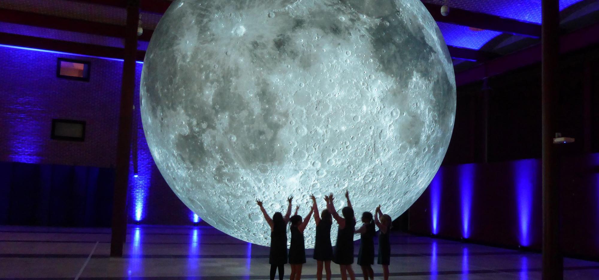 A large moon sits centrally in a room with a group of children reaching out to touch it