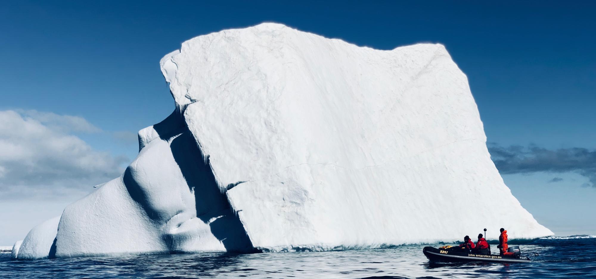 A large white iceberg is framed against a brilliant blue sky and clear rippling blue water, with a tiny boat of researchers at the bottom right corner of the ice berg showing its sheer scale