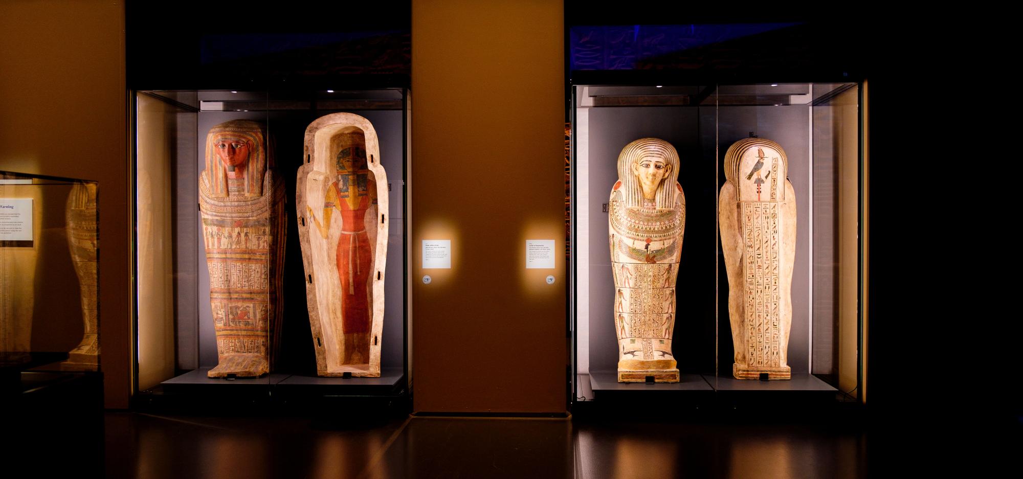 A dramatically-lit gallery with two coffins standing upright on display. The coffin on the left shows an image of an Egyptian goddess while the coffin on the right shows intricate hieroglyphs.