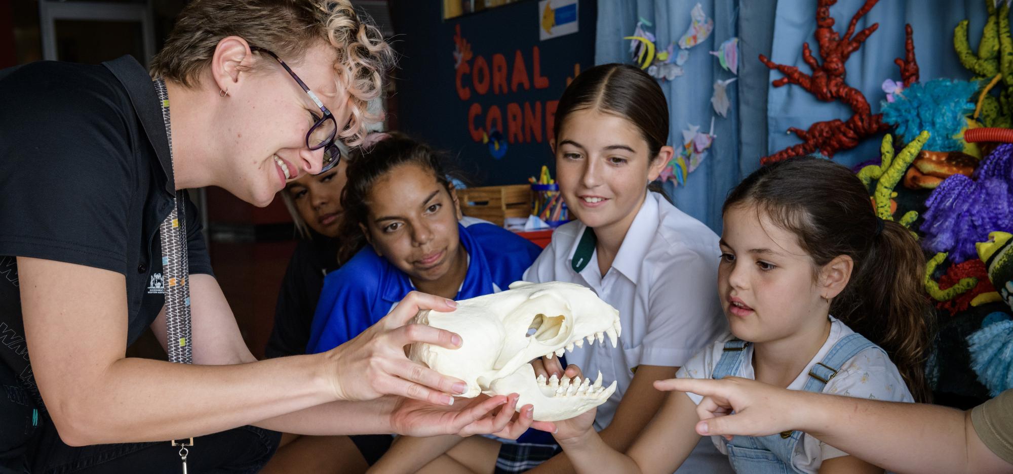 A person with short curly blonde hair and black glasses wears a black Museum uniform and smiles as they hold a white skull model up for school children to look at. The children sit cross legged and touch and point to the skull as though with interest