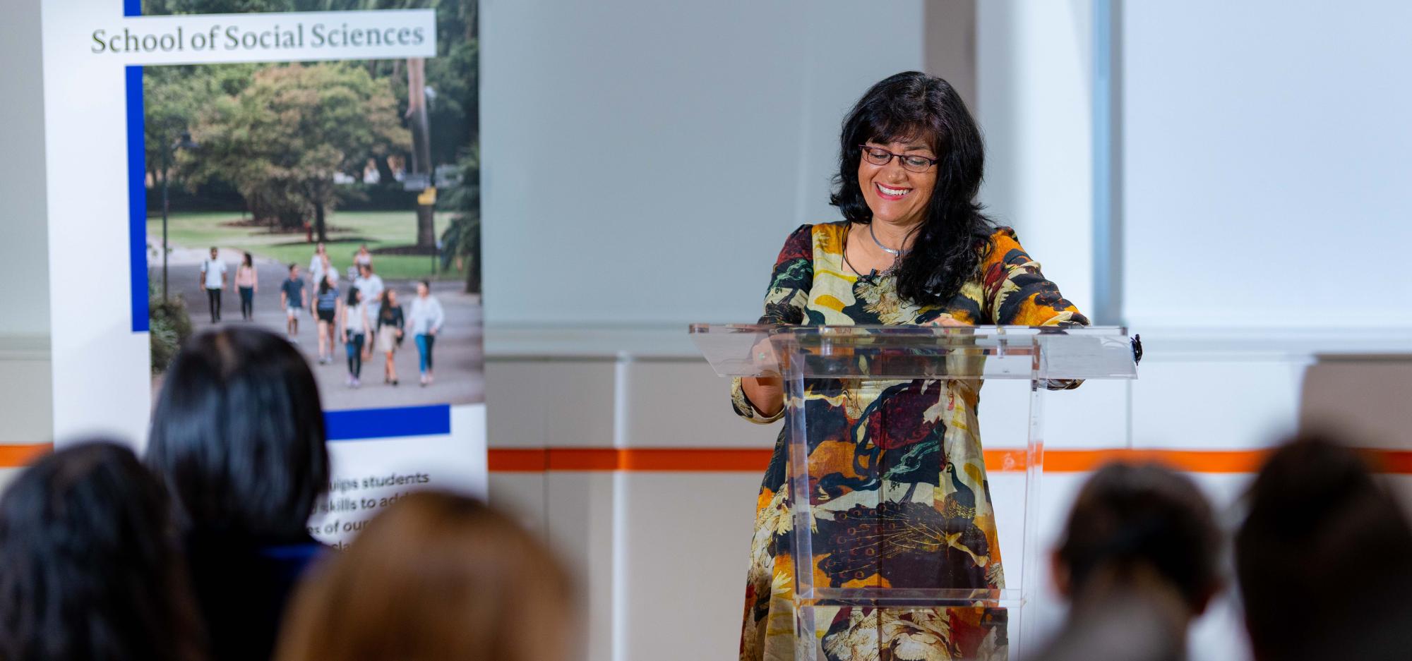 Pilar stands at a lectern in front of a UWA banner in a white conference room wearing a vivid autumnal patterned dress with her long black hair over her shoulder. She smiles as though in mid-sentence while she presents to a room of people.