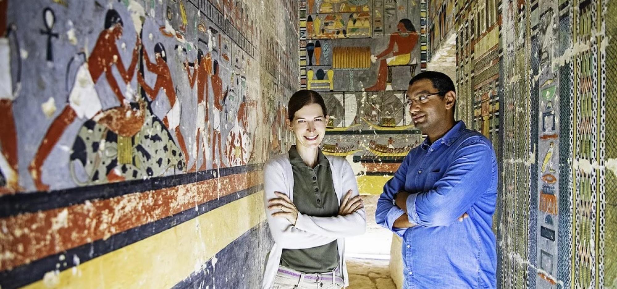 Dr. Mohamed Megahed and Dr. Hana Vymazalová stand side by side with their arms folded, facing the camera. They are situated inside an Egyptian tomb adorned with illustrations on the walls.