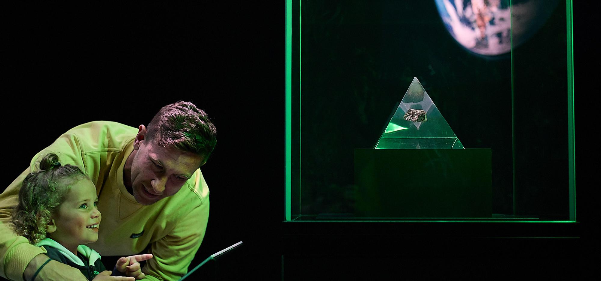 An adult and a young child with curly hair are bathed in green light from a Museum exhibition as they stare with interest at a showcase containing a glass pyramid holding a small section of moon rock.