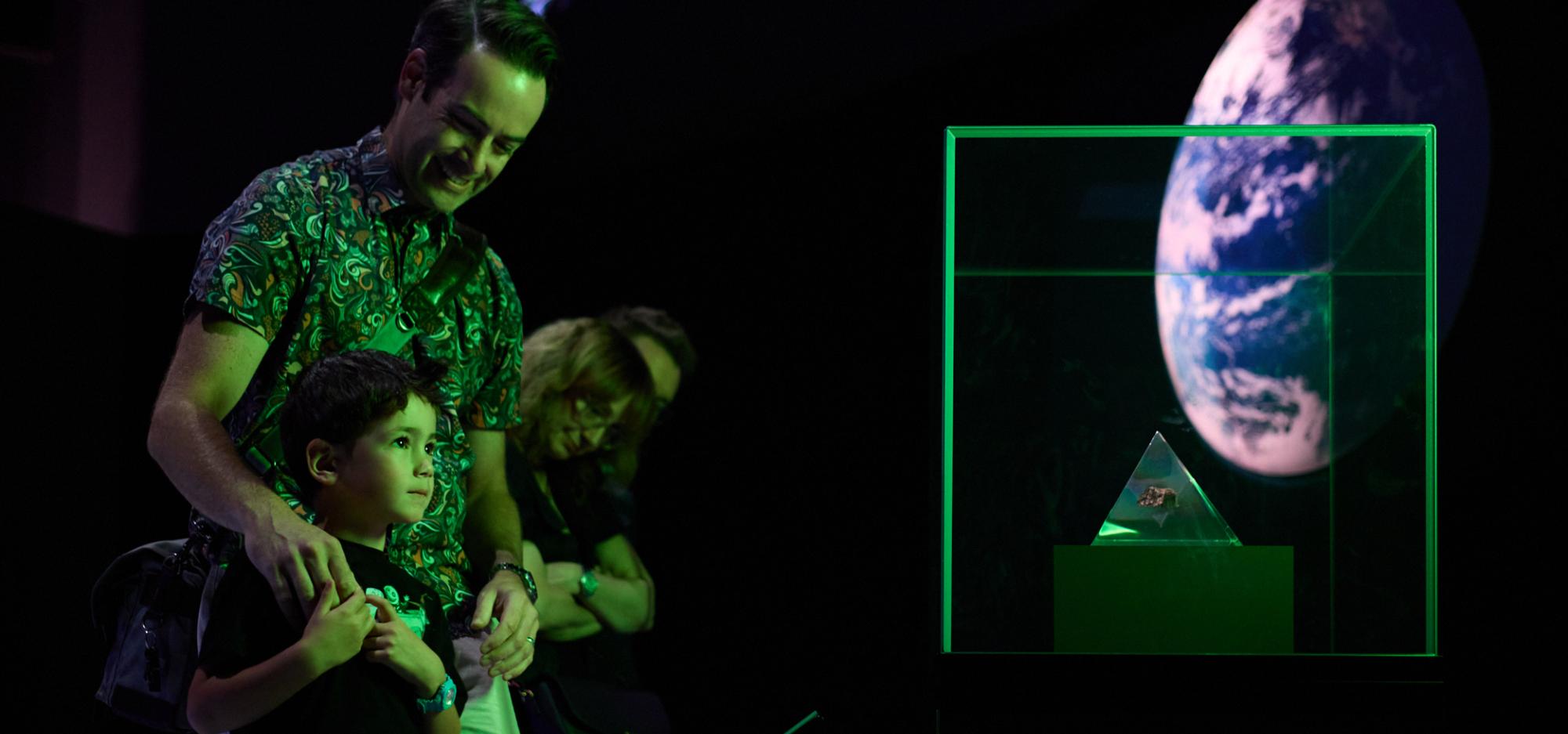 An adult and a young child are bathed in green light from a dramatically lit Museum exhibition. The adult smiles and looks down at the child as the child stares with interest at a glass pyramid containing a moon rock sample in a showcase.