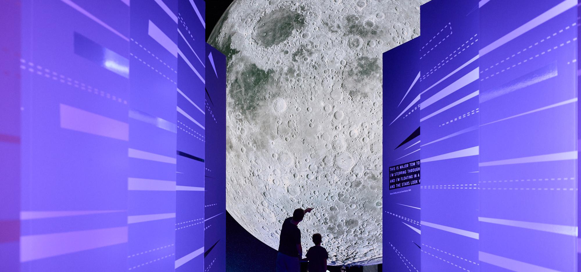 A large moon sculpture is visible down a corridor of brightly lit purple walls designed in a sci-fi aesthetic style with two people who appear small in comparison taking a selfie while in silhouette 