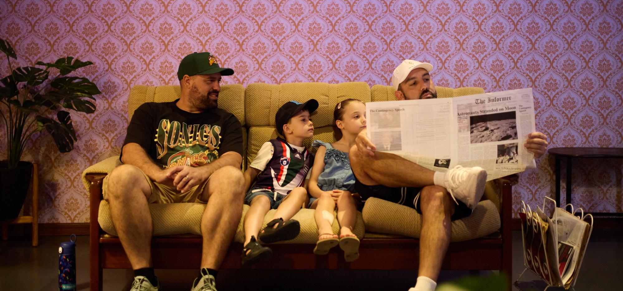 Two adults and two young children sit on a 1970s-style couch and read a newspaper in a Museum gallery.