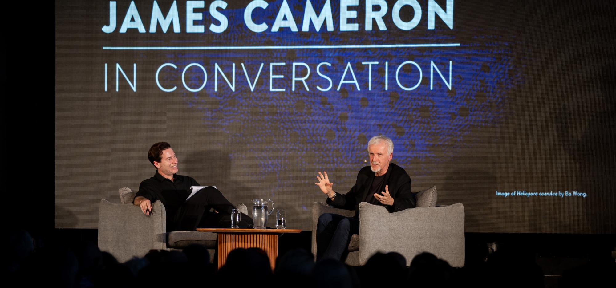 James Cameron dressed in black with grey hair sits in a grey chair and gestures passionately as he speaks at a large crowd. The words James Cameron In Conversation are projected behind him and Ben Shea, also dressed in black with dark hair reclines with a smile on his face in a grey chair opposite.