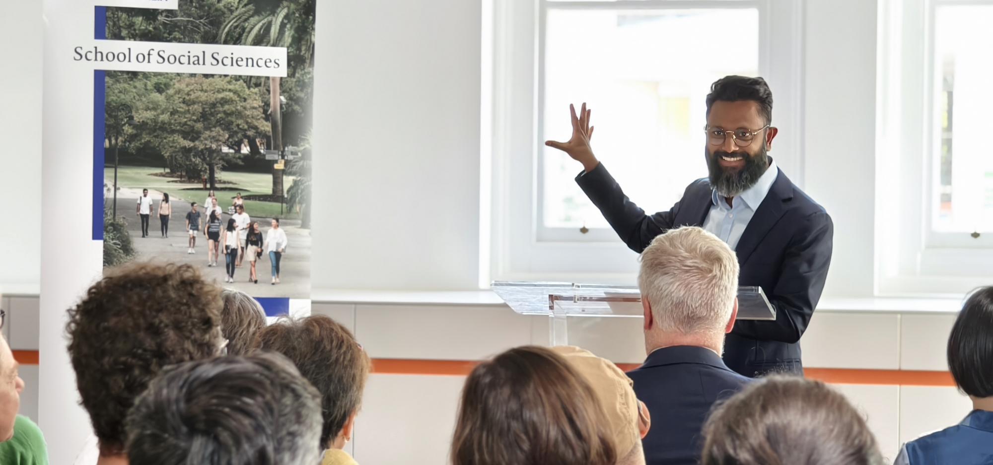 James Jegasothy wears a black suit and light blue shirt and thin wire frame glasses and gestures passionately as though in mid presentation. They stand at a lectern in a brightly lit space, in front of an audience of people.