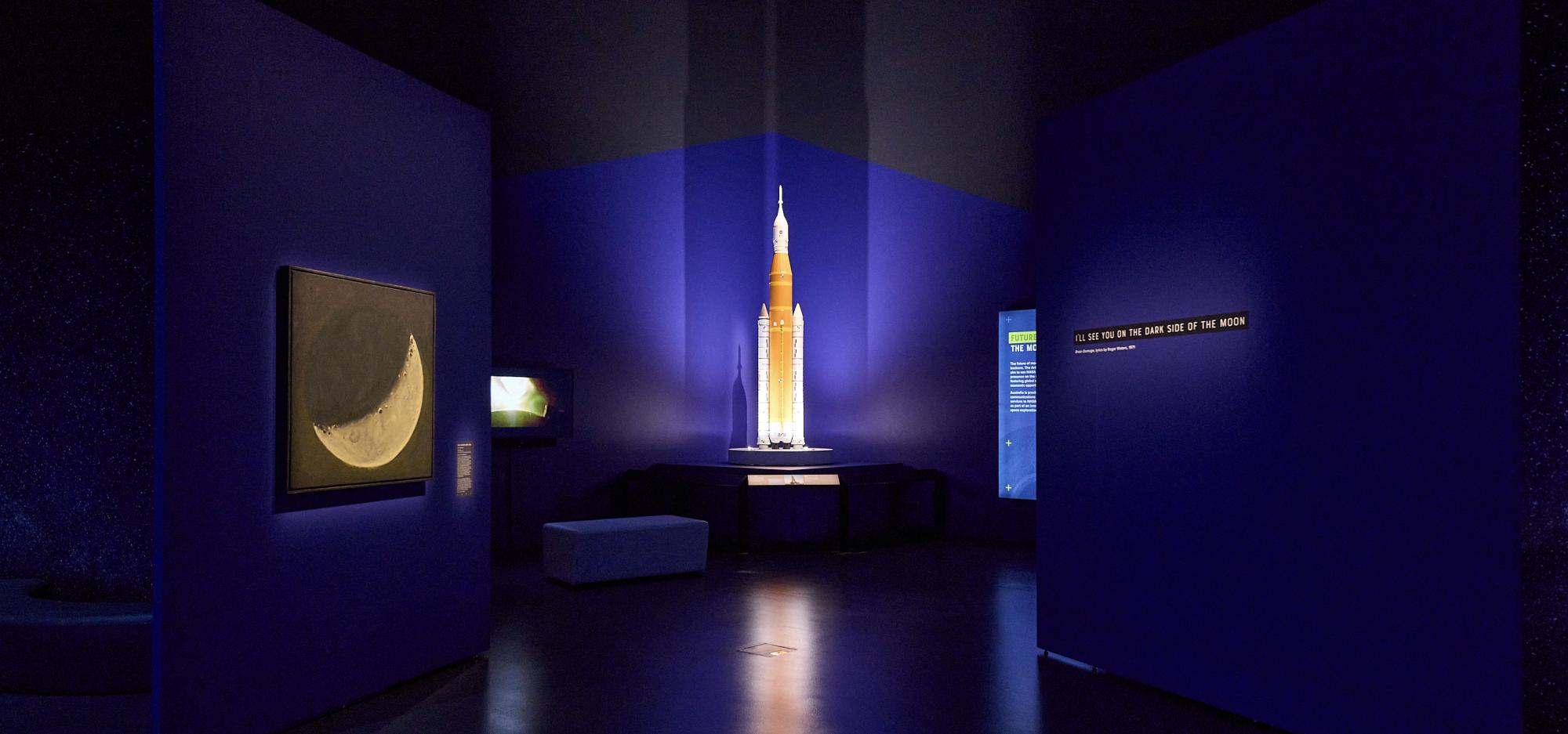 The walls of a dramatically lit Museum gallery bathed in blue light open to reveal a brightly lit rocket replica on a small plinth.