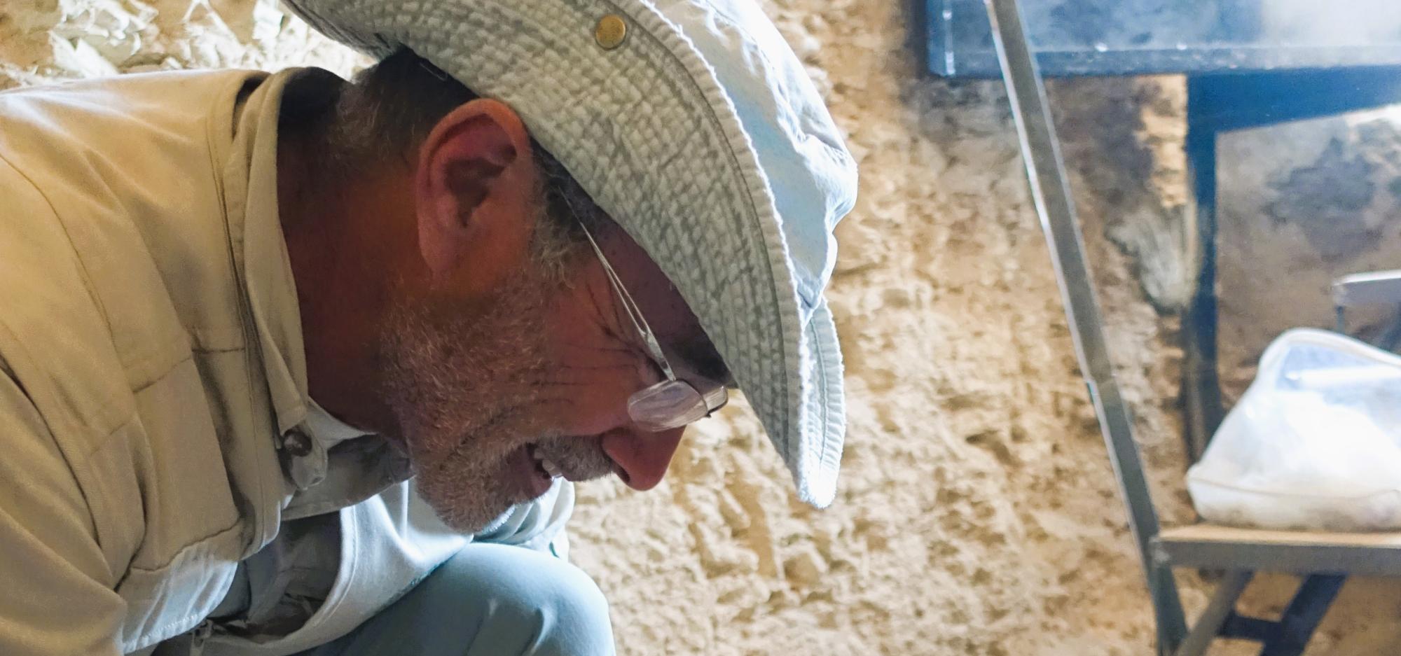 Man wearing hat and glasses crouched over ancient object brushing away dirt with paintbrush