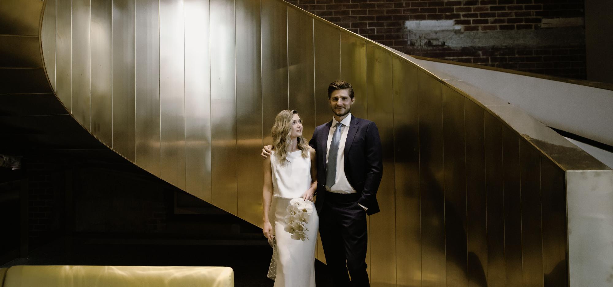 A couple in wedding attire stand beside a curving golden staircase
