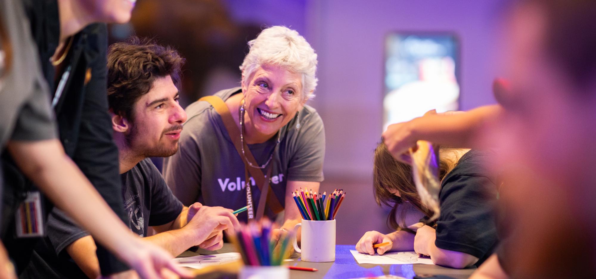 A small group of people sit at a table with paper and pencils in a colourful purple and blue lit gallery. A volunteer in a grey shirt leans over the table and smiles at the group