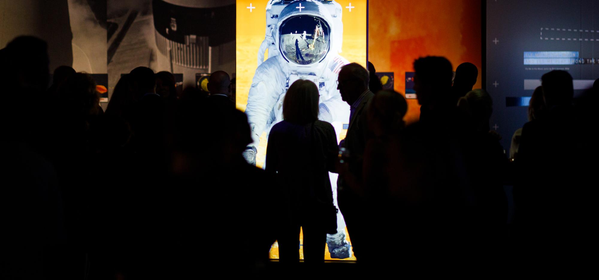 A brightly lit tall image of an astronaut in a darkened gallery surrounded by a large amount of silhouettes of people