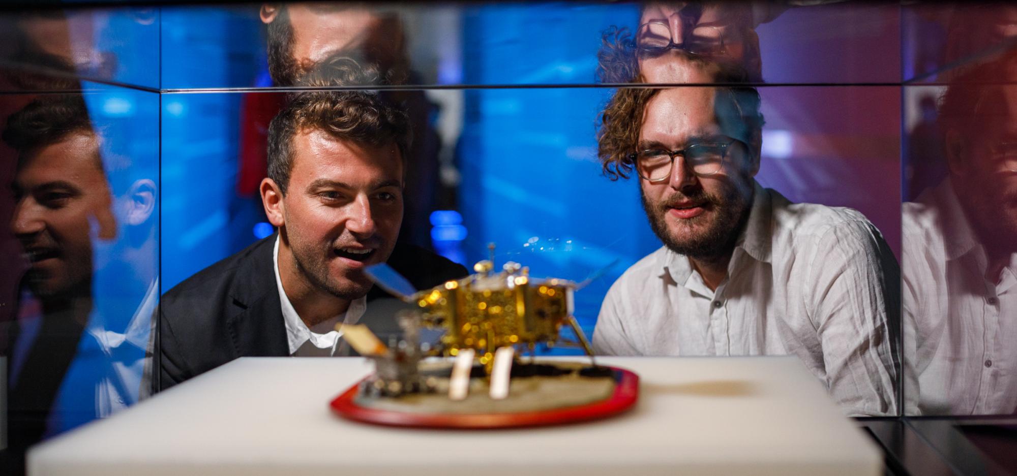 A person wearing a black suit jacket with short-cropped curly hair and a person with a white button up shirt, short hair and thin rimmed glasses stare with expressions of interest at a small gold spacecraft model in a glass Museum showcase