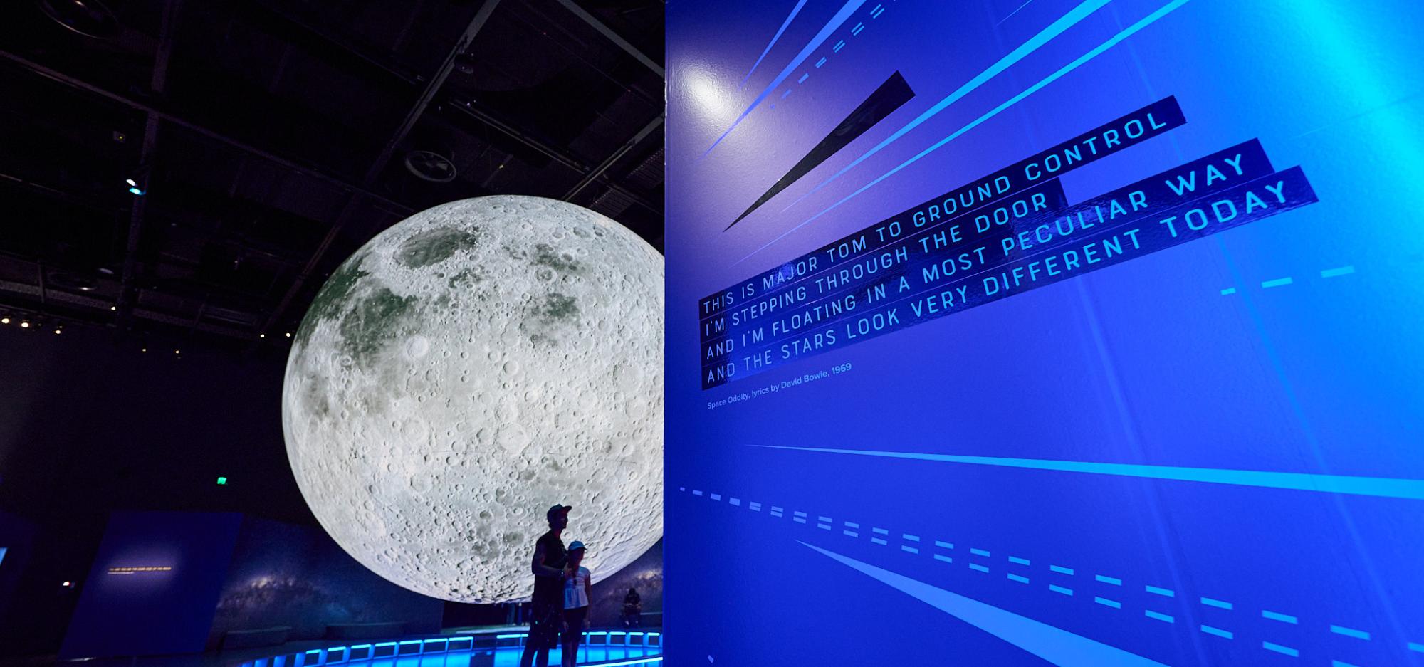 A dramatically lit large moon sculpture hangs in a dark gallery partially obstructed by a wall bathed in blue light with the lyrics to David Bowie's Space Oddity in black and white text