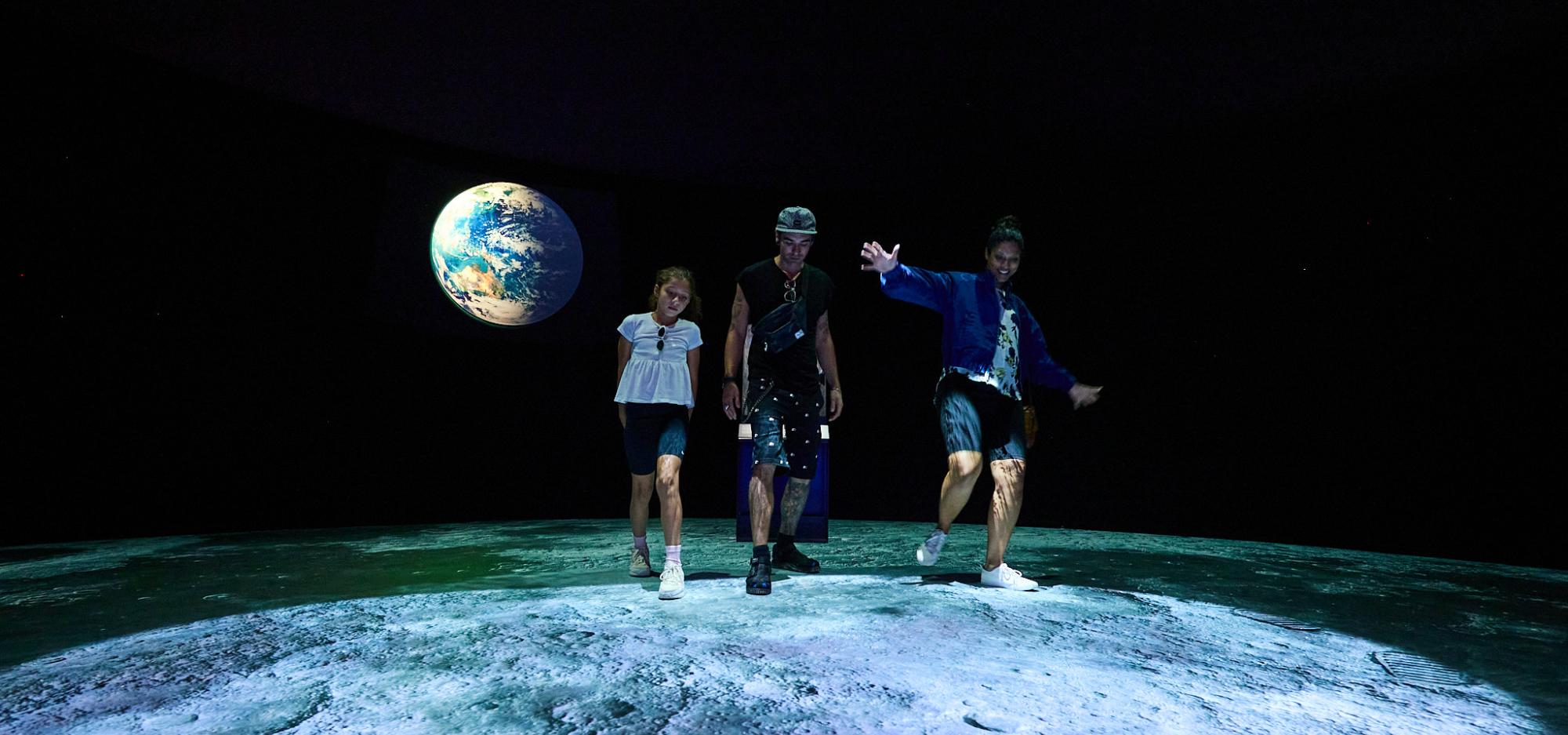Three people walk over a projected image of the moon surface with footsteps. The installation sits in a darkened Museum gallery with a projection of the earth on the back wall.