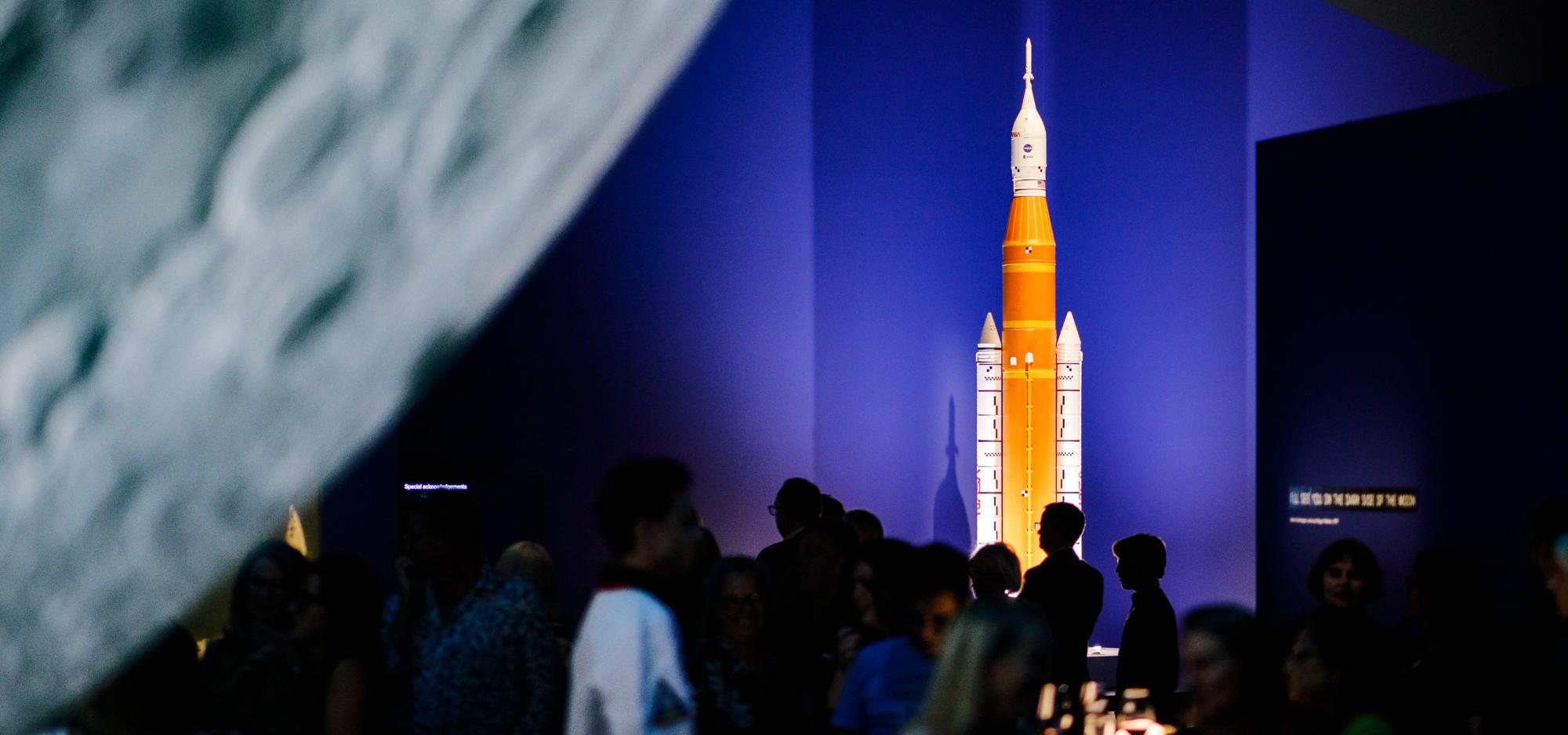 A orange and white model of a rocket is brightly lit in a darkened Museum space. A section of a large moon sculpture shines in the left hand side of the frame and silhouettes of people mill around beneath it.