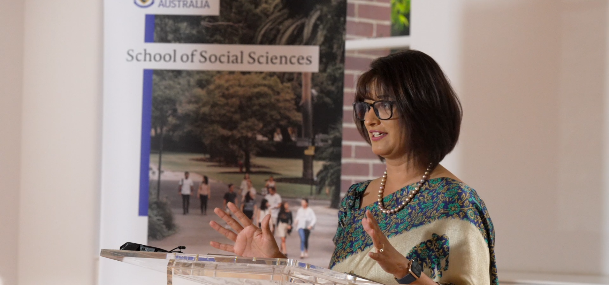 Dr Parwinder Kaur has short brown hair and black glasses and wears a blue and white shawl and a pearl necklace. They stand at a lectern in front of a UWA social sciences poster and gesture with their hands mid-presentation.