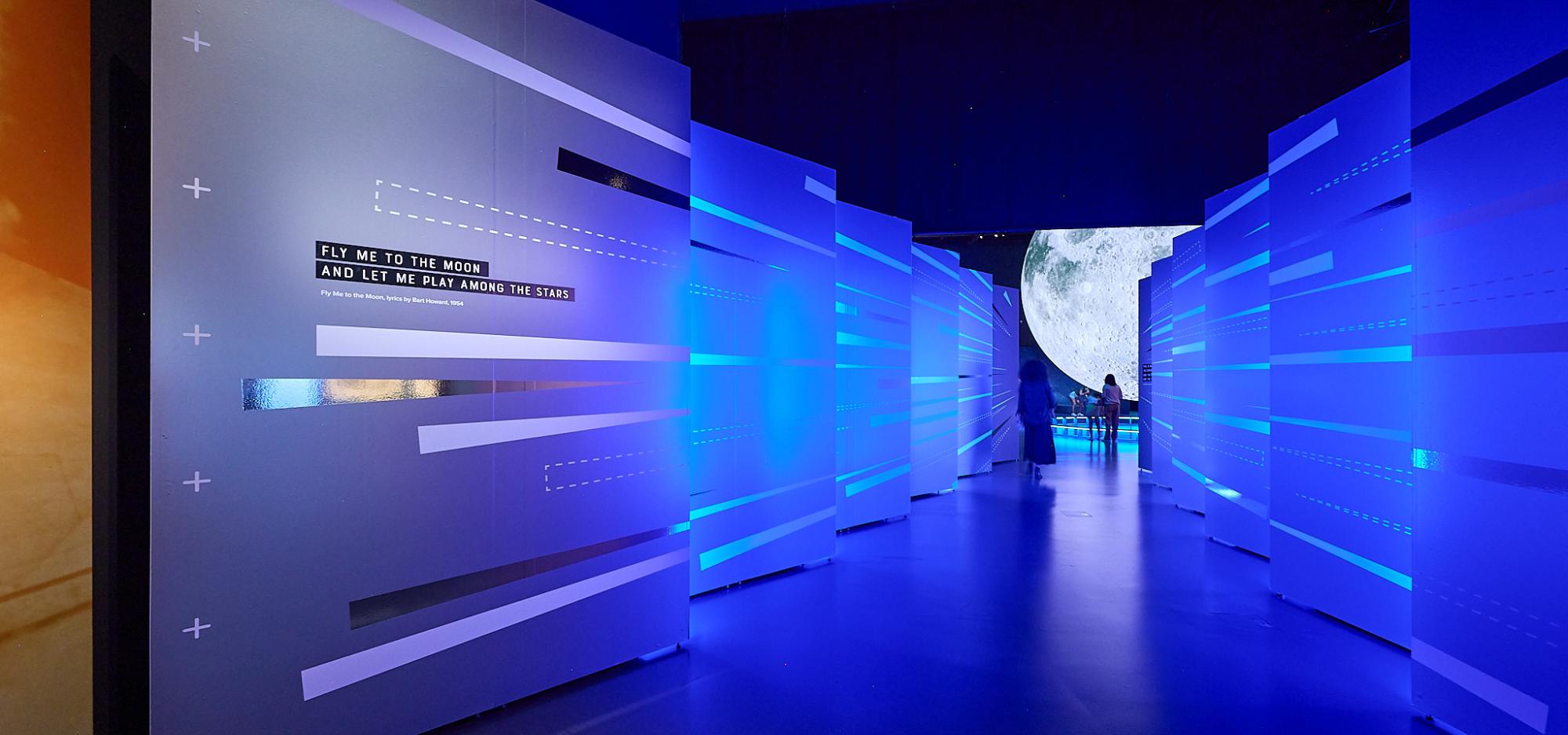 A hallway illuminated by coloured lights with reflective graphics leading to a large moon display.