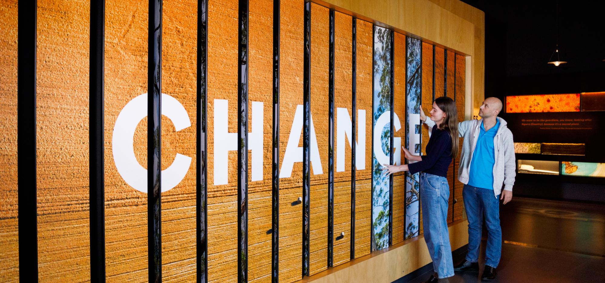 Two people interact with a wooden panel displaying the word "CHANGES" in large white letters.
