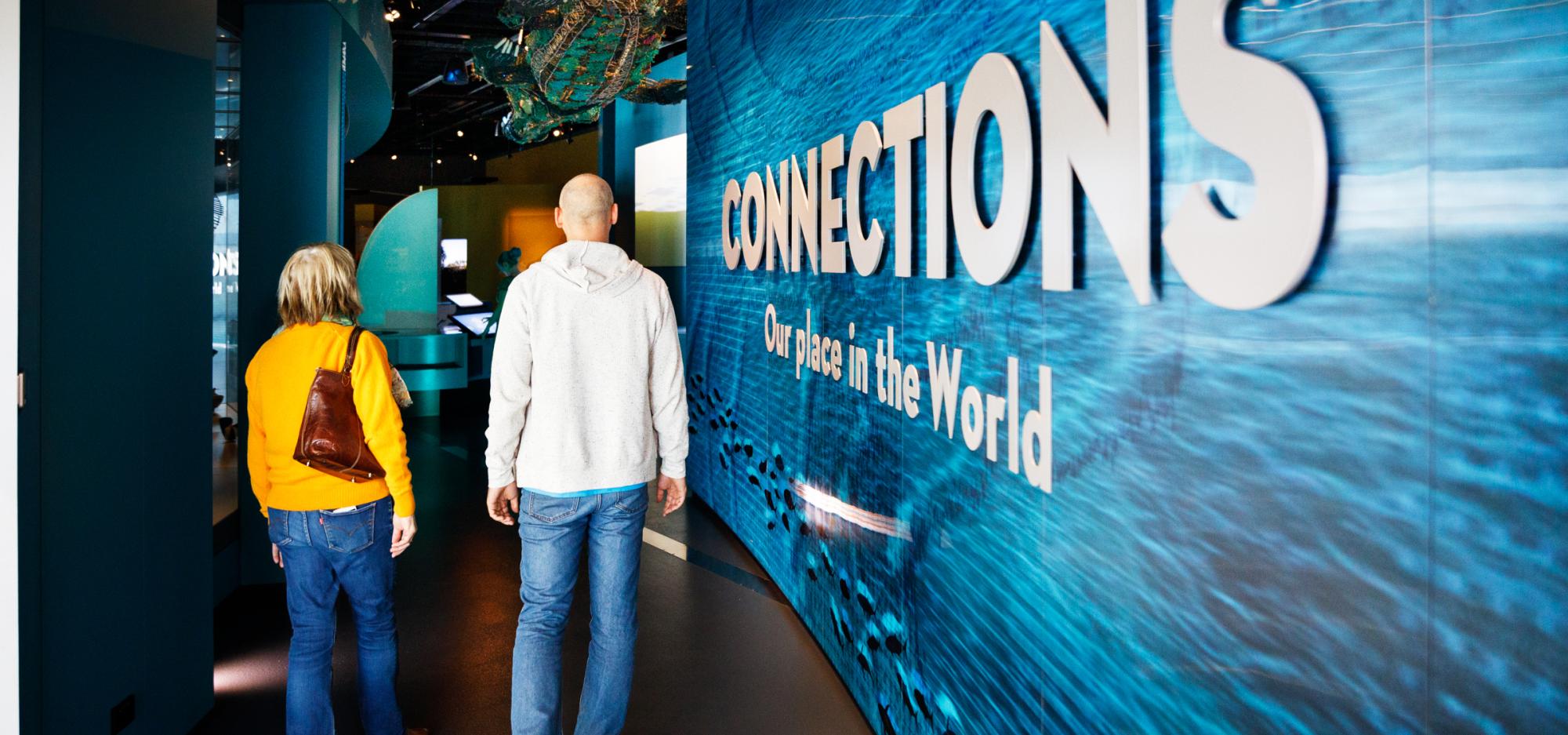  Two people walk through a museum exhibit with a large blue wall displaying "CONNECTIONS Our place in the World."