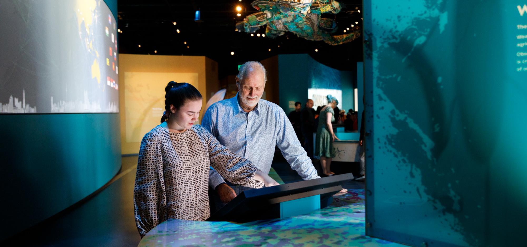 People interacting with a touchscreen exhibit in a modern, dimly-lit exhibition space with a large model creature suspended above.