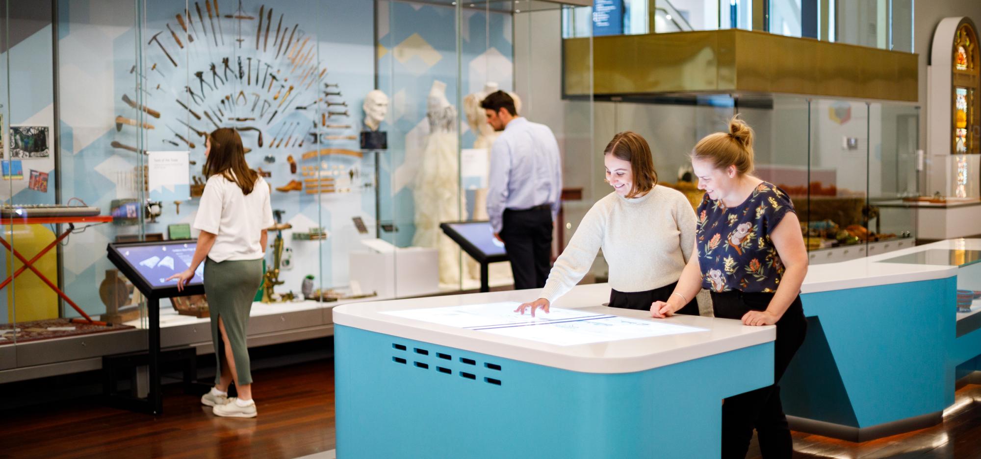 Visitors interact with exhibits in a modern museum, including digital tables and display cases.
