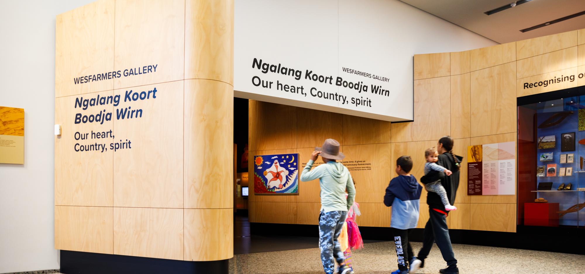 Entrance to Wesfarmers Gallery with wood paneling and text 'Ngalang Koort Boodja Wirn: Our heart, Country, spirit.' Five visitors approach the entrance.