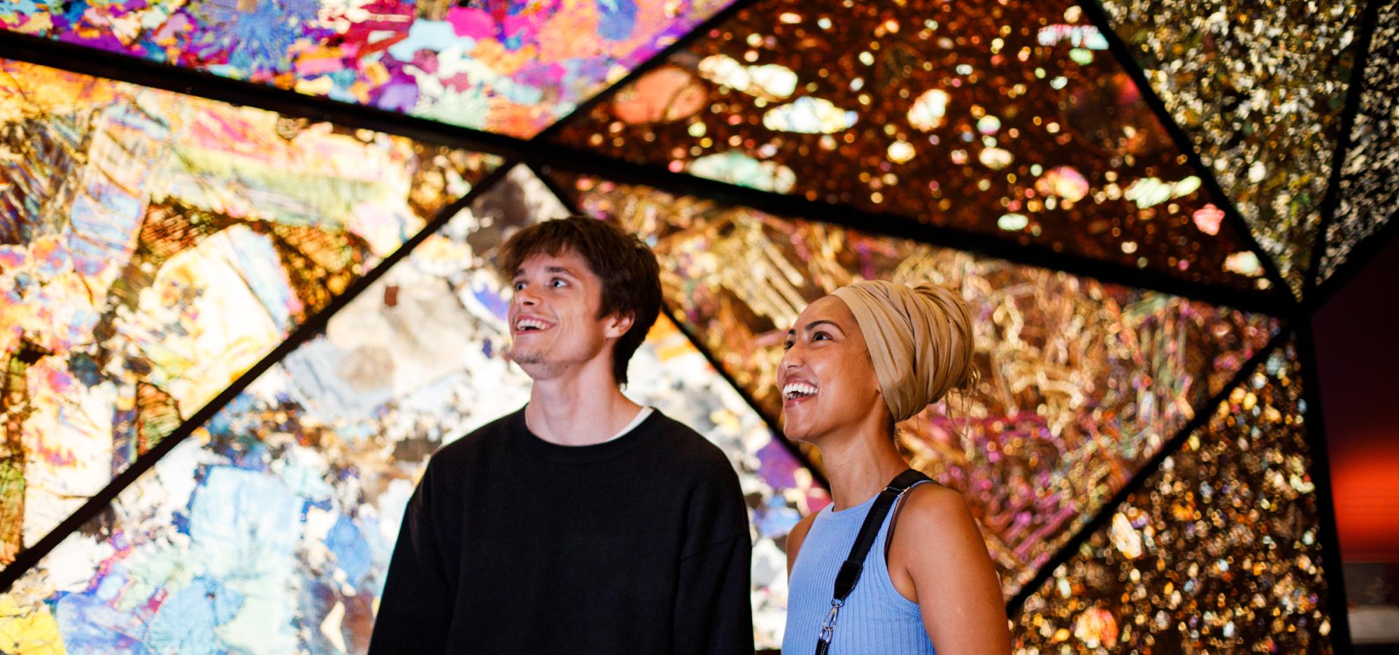 Two people smiling and looking upwards in a colorful, geometric, and illuminated space with intricate patterns.