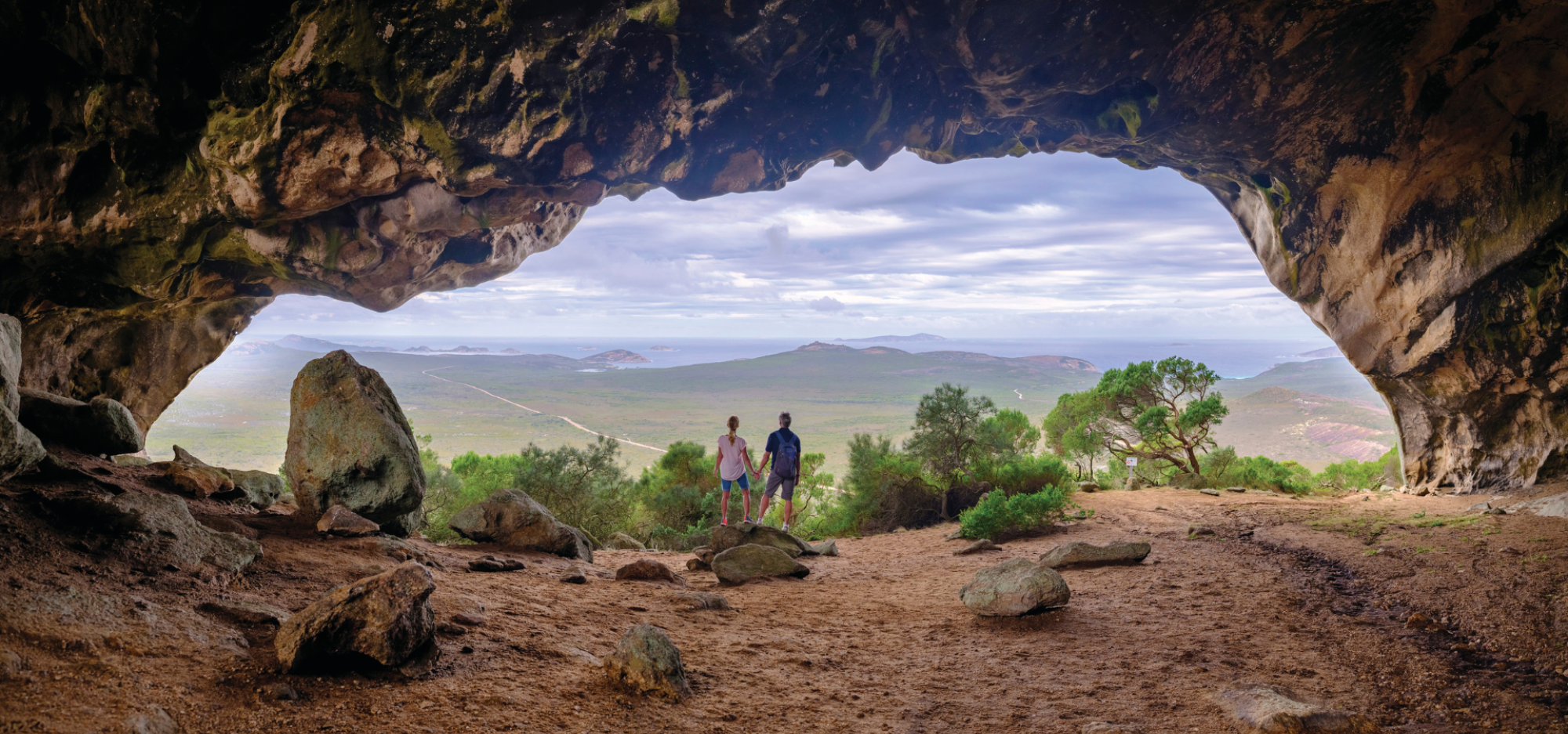 A wide angled shot of two people holding hands with their back to the camera, standing in the mouth of a large cavern. A green landscape stretches out before and beneath them