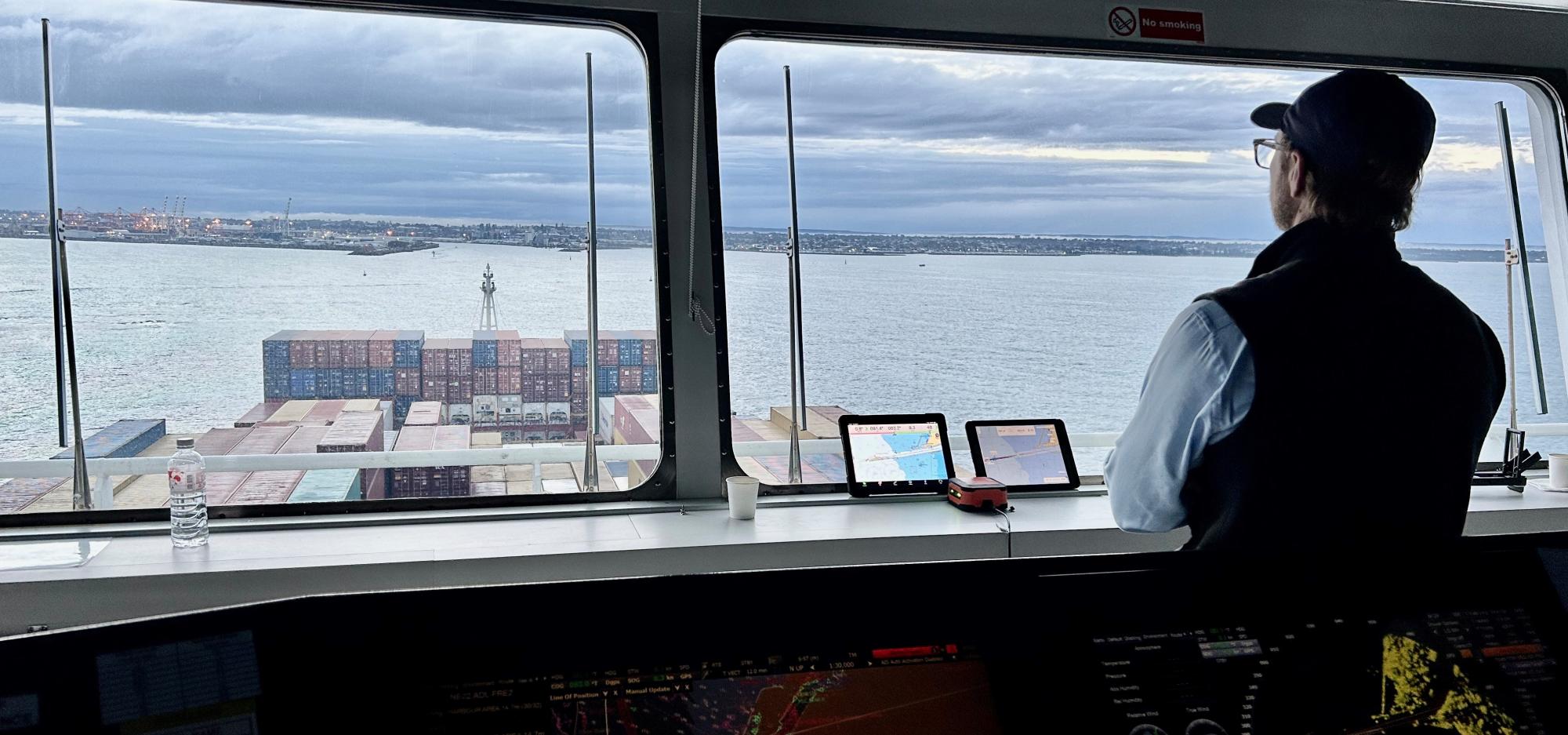 an adult person in slight silhouette stands inside a ship indoor viewing deck with controls. They look out through the large window onto a harbour with sea containers