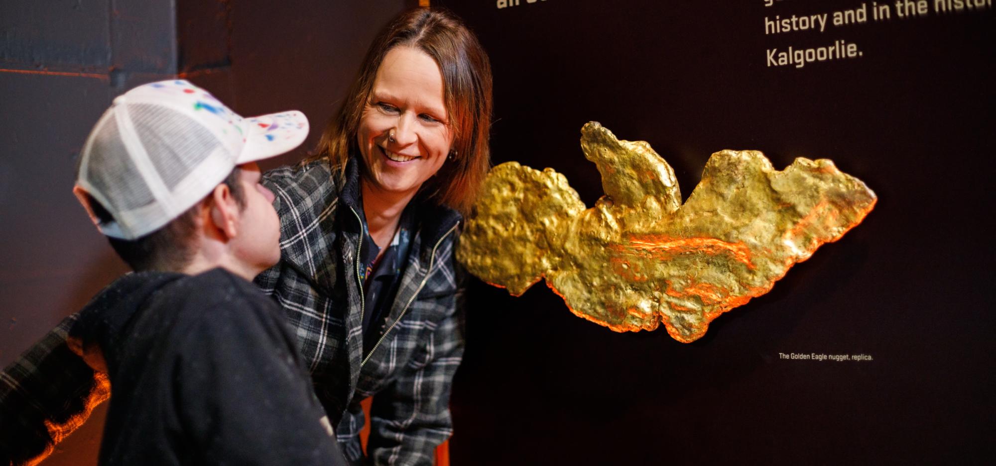 Smiling woman and young boy standing in front of a black graphic panel showcasing a large nugget of gold