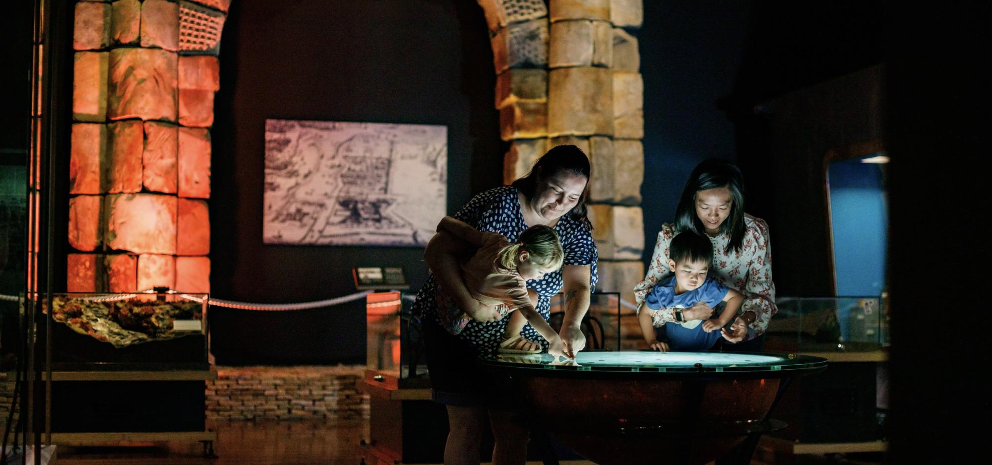 Two people each hold a young child who interact with a brightly lit display table in a dimly lit museum room featuring large stone arches and a framed artwork on the wall.
