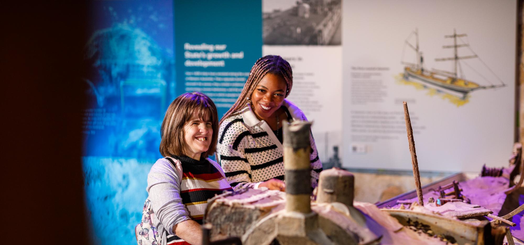 Two people observe a detailed shipwreck model at the WA Maritime Museum with a series of colourful panels depicting ships and shipwrecks displayed behind them