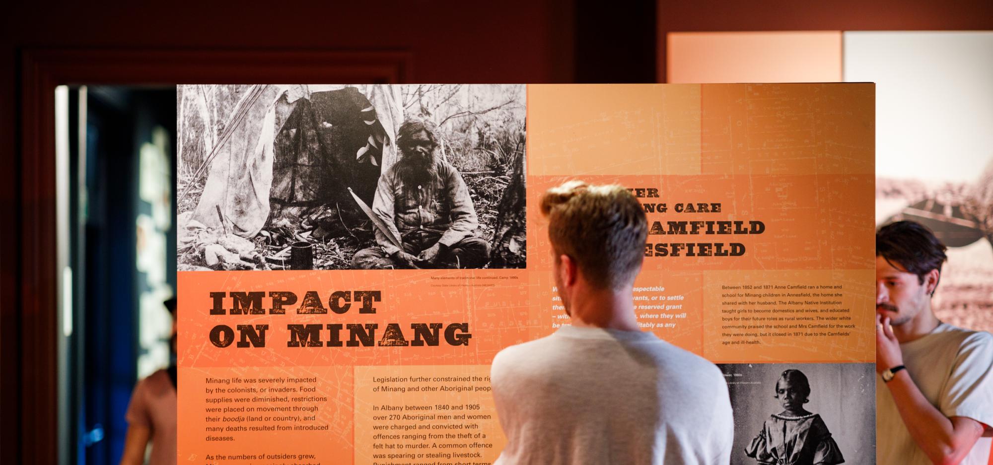 Man reading a graphic panel describing impacts of colonists on the Minang Aboriginal people with two black and white images of an elderly Minang man sat outside a tent and young Minang girl in a Victorian era dress holding a hat 