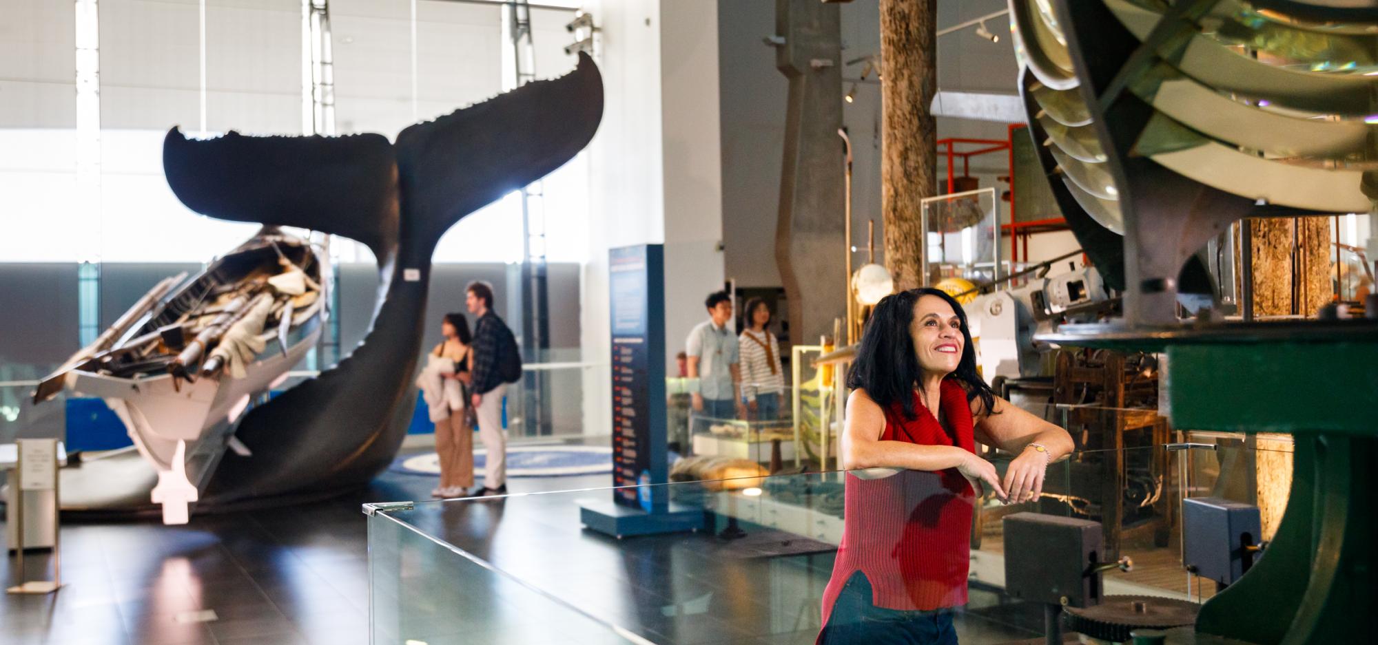 Woman smiling and leaning on a glass case in a museum, with large historical maritime equipment and a whale tail sculpture in the background.