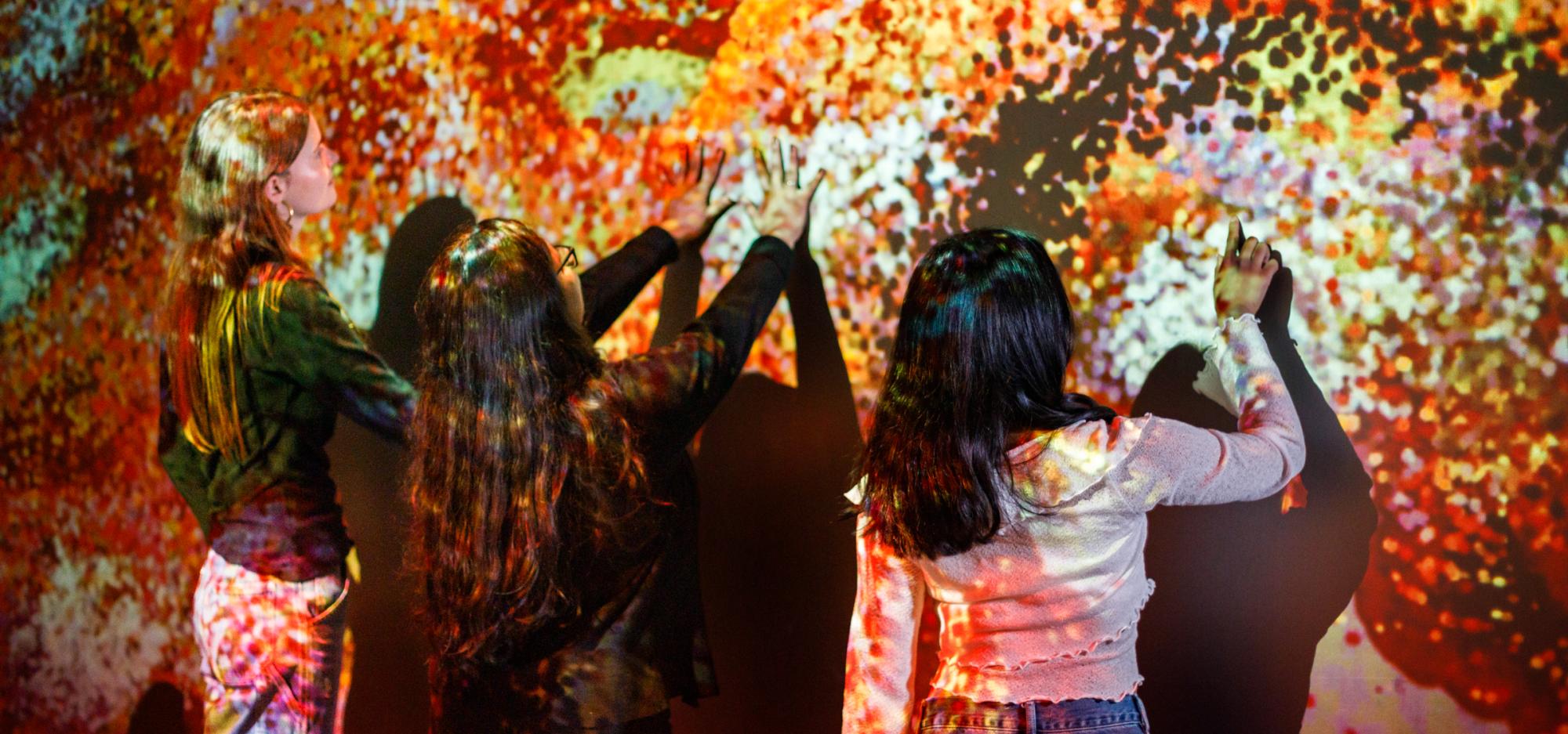 Three people interact with a colourful projection of Aboriginal artwork on a wall.