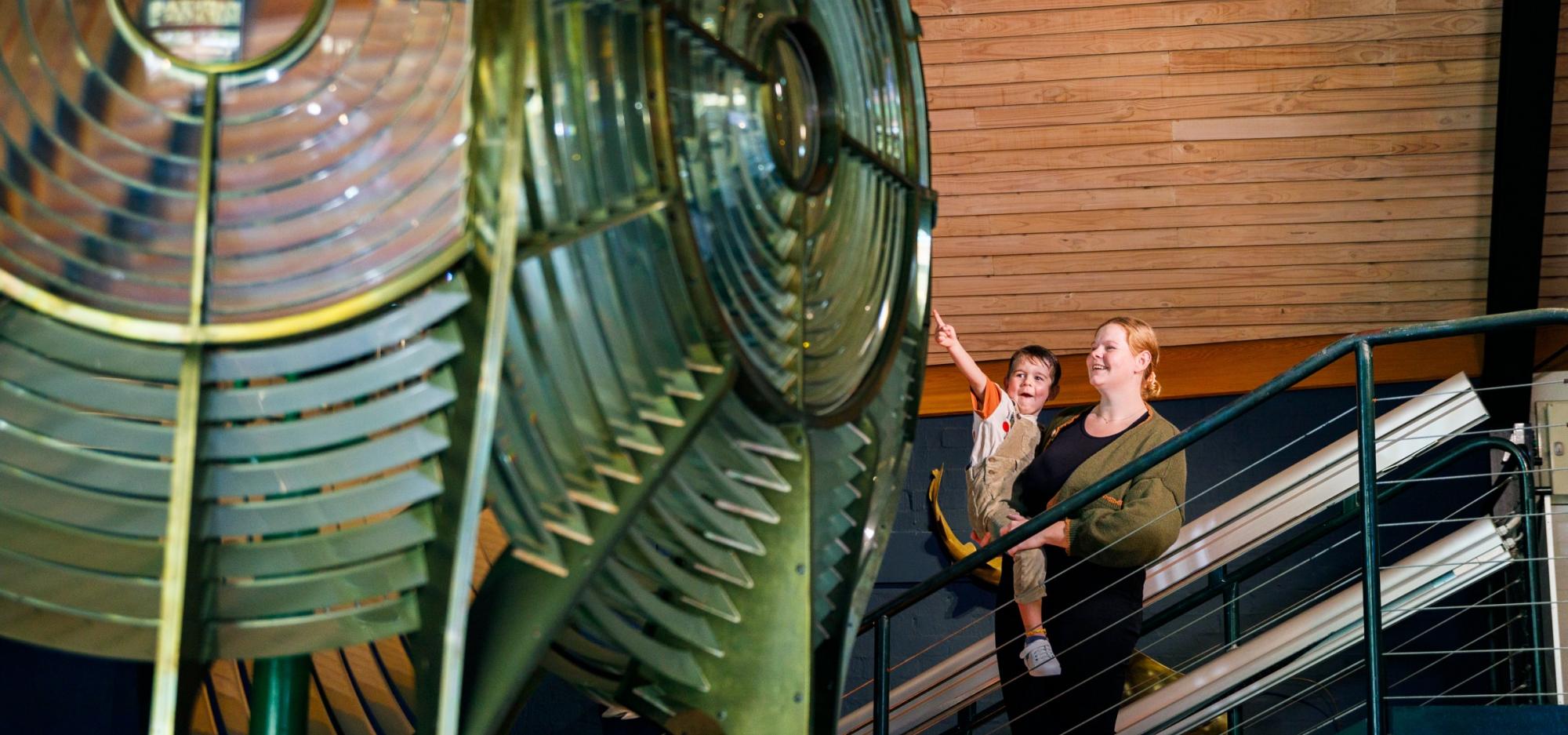 An adult holding a small child who is pointing with excitement at a large green lighthouse lamp