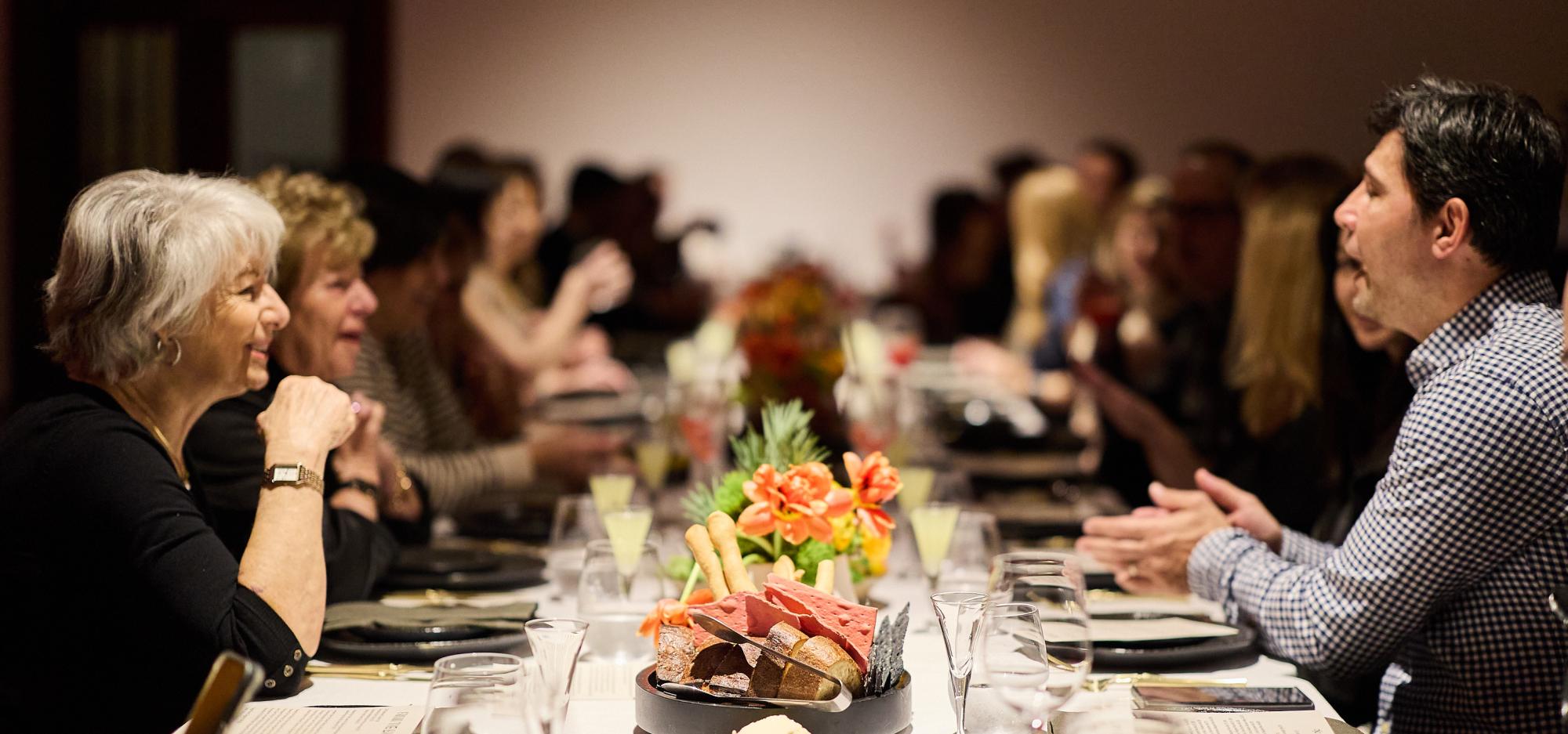 People conversing around a long dining table with a centerpiece of bread and orange flowers.