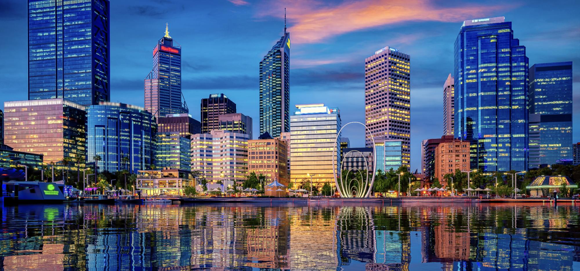A wide shot of Perth city taken from across a river at night with buildings of varying heights brightly lit by yellow office lights reflecting blue orange and yellow lights off the river and backlit by a blue sky with bands of orange clouds