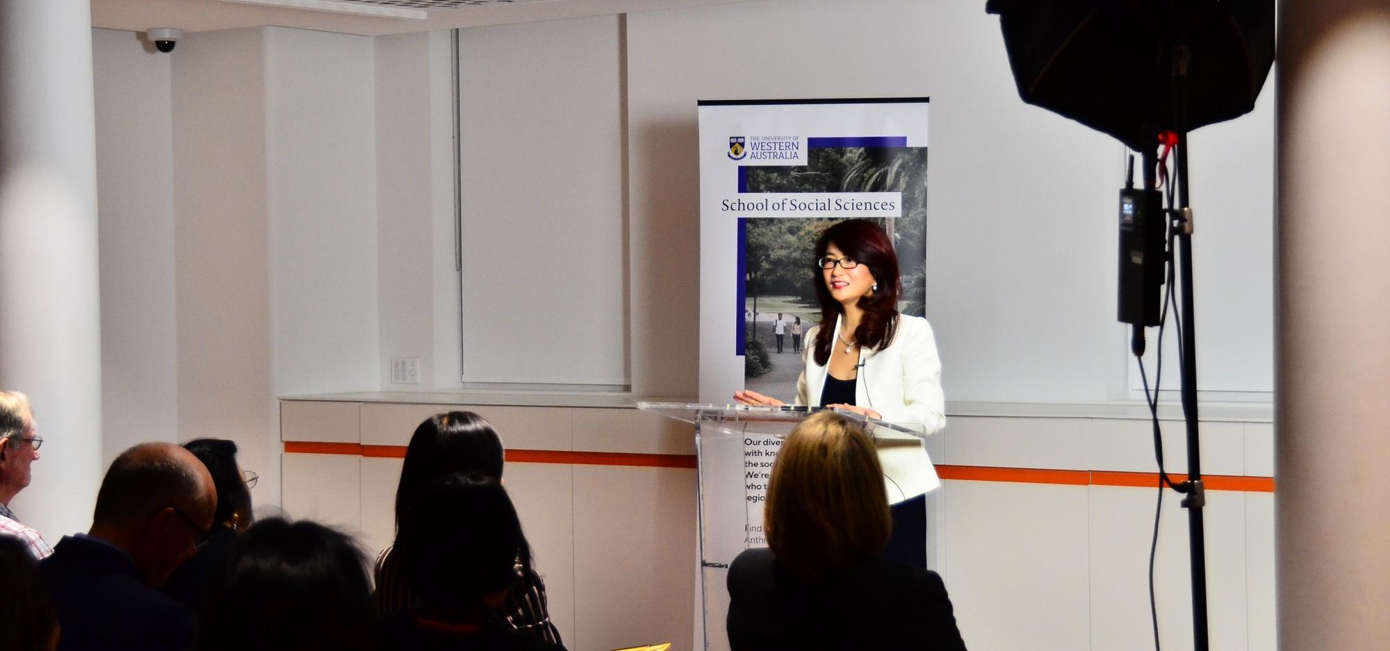 A person in a white jacket stands at a lectern in a white room and presents to a seated audience in front of them.