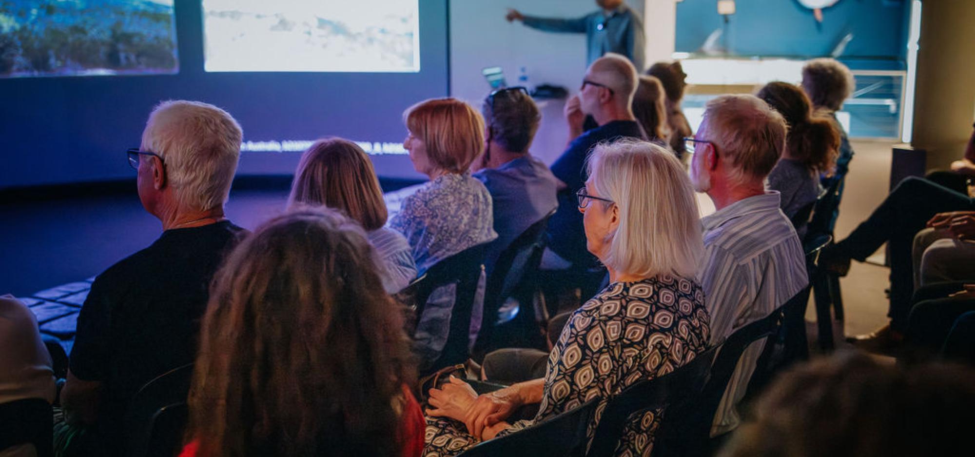 people seated facing a large curved screen listening to a person in the background near the screen talking 