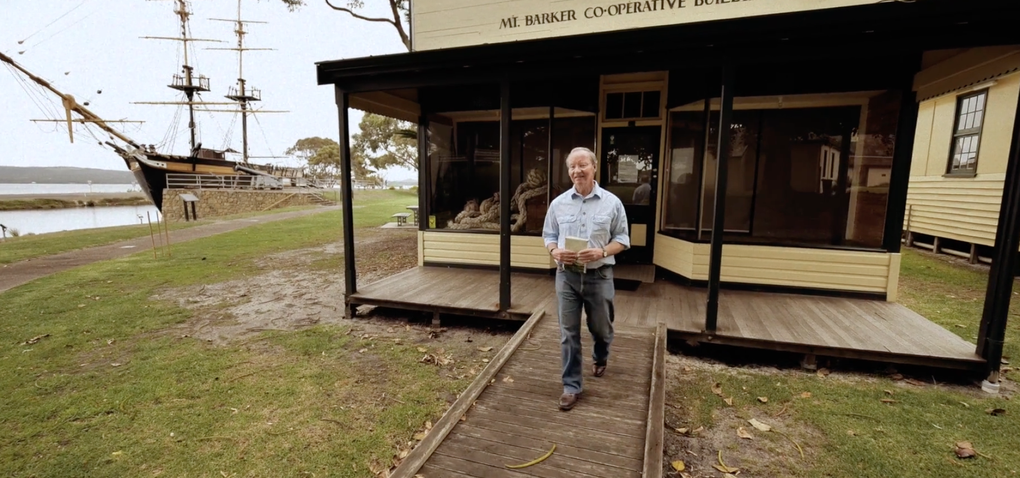 Person walking from the Mt Barker Co‑operative Building, with a historic sailing ship and waterfront in the background.