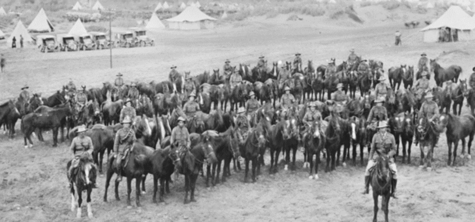 Black-and-white photo of mounted troops lined up on horseback, with tents of a military camp in the background.
